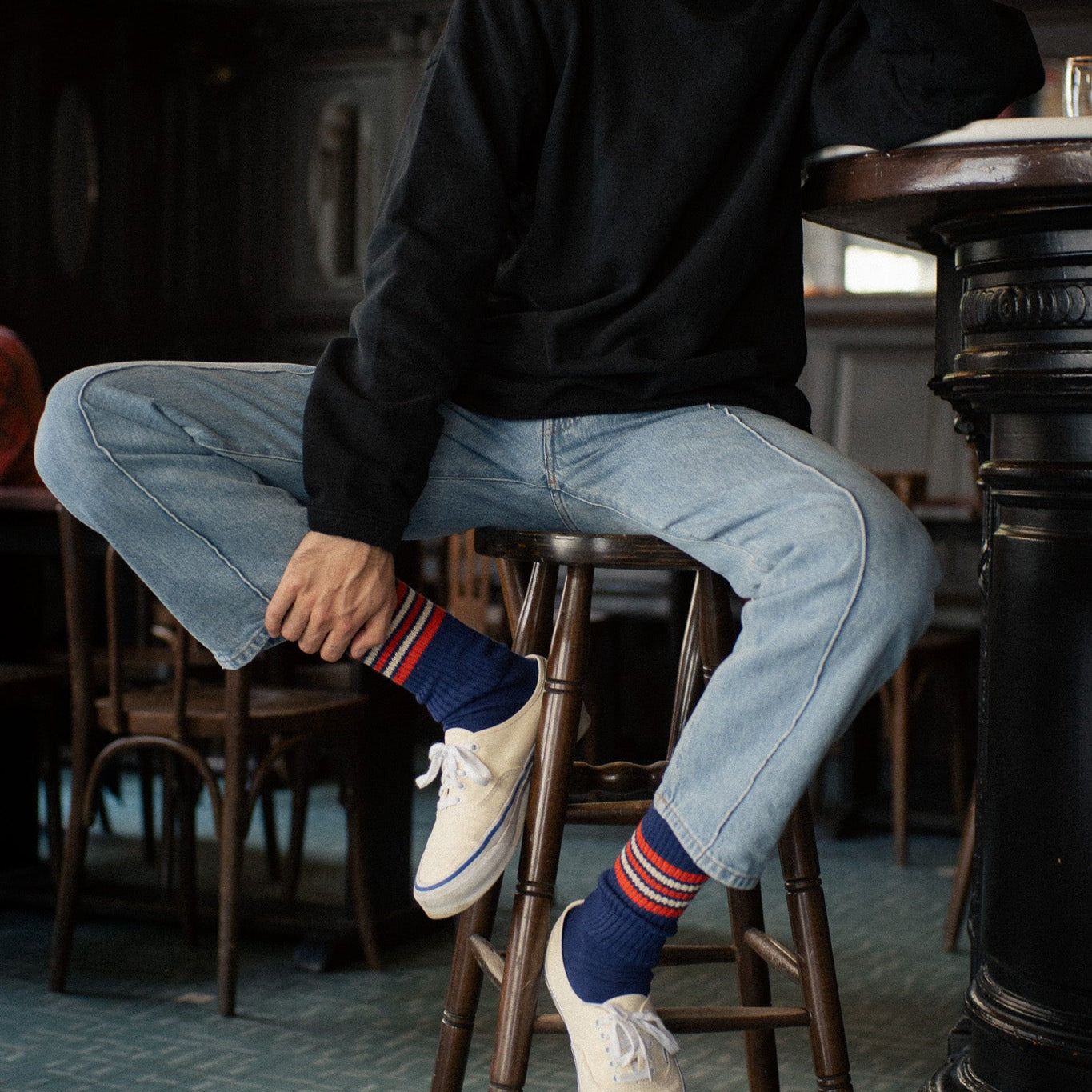 Escuyer blue, orange and ecru 5 Stripes socks with white canvas sneakers and jeans, seated on a bar stool
