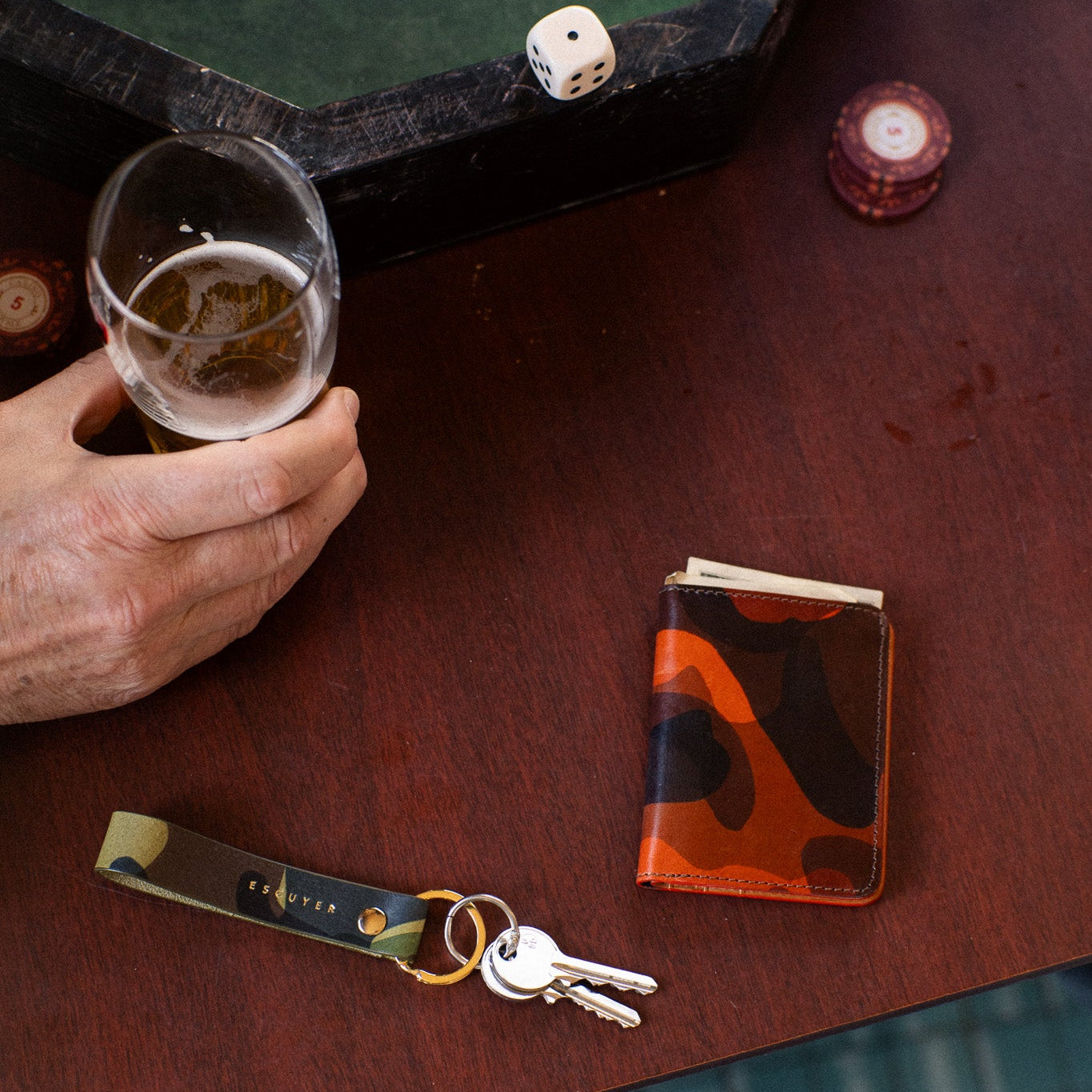 Escuyer slim wallet in hand-dyed orange camouflage leather on a bar table beside a light green camouflage keychain