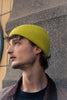 Man wearing a yellow knit beanie against a textured wall.