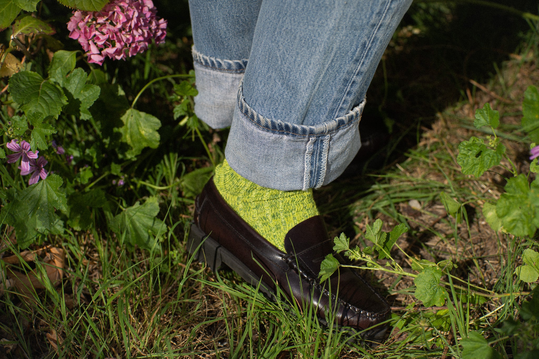 Person wearing green socks and black shoes with rolled-up jeans standing in a garden.