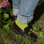 Person wearing green socks and black shoes with rolled-up jeans standing in a garden.