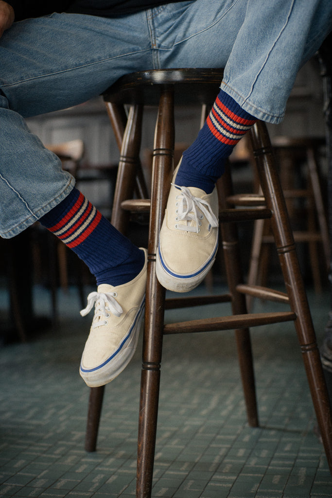 Person wearing blue jeans and white sneakers from Vans Skateboards with red and blue striped socks from Escuyer brand