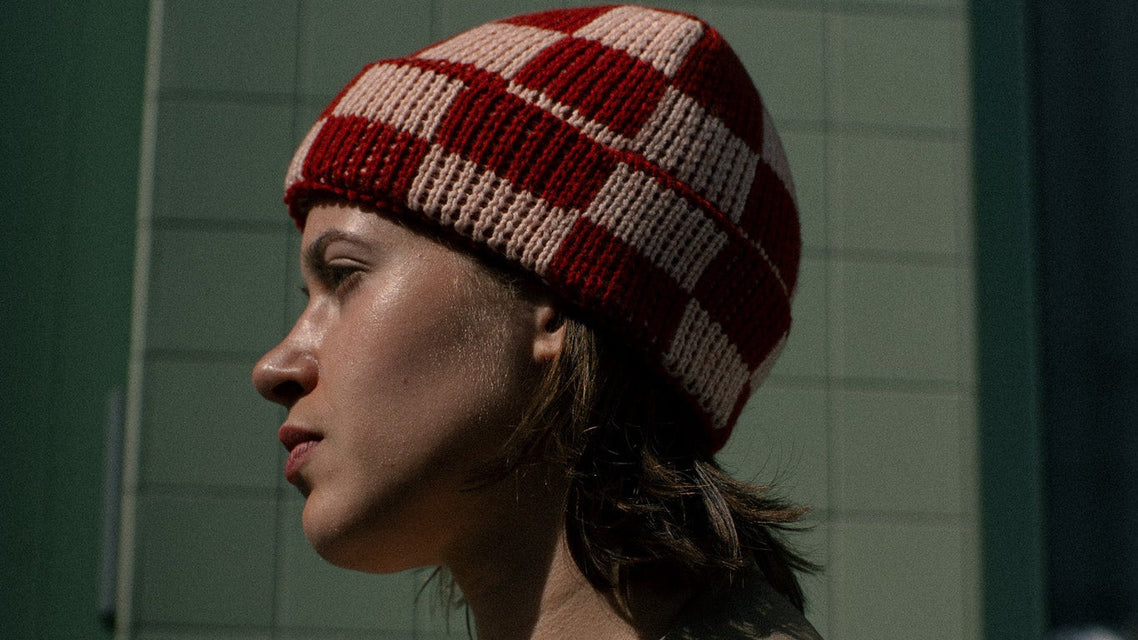 Person wearing a red and white checkered beanie and brown coat against a green tiled wall.