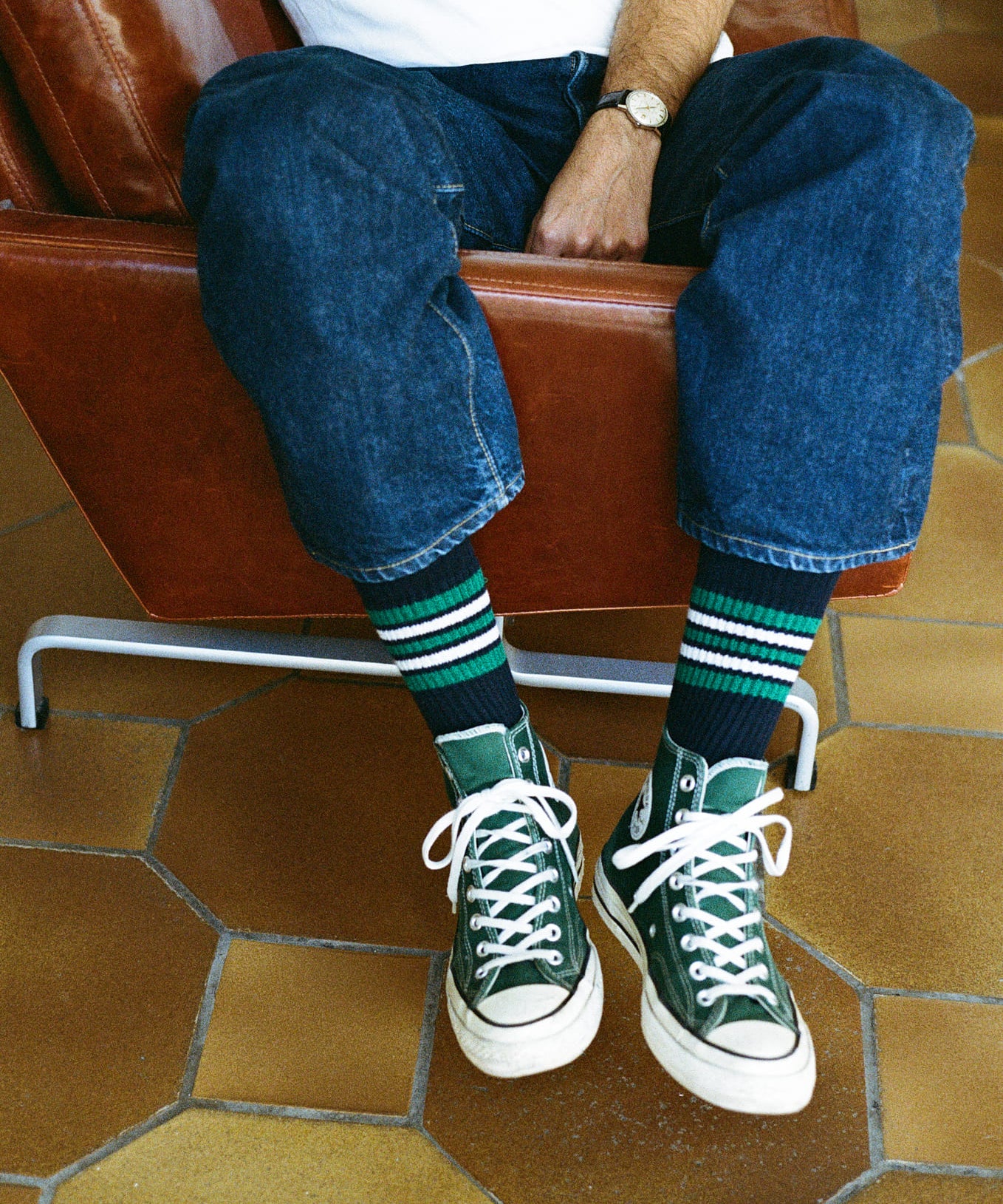 Person wearing green sneakers and blue jeans sitting on a brown leather chair with a tiled floor.
